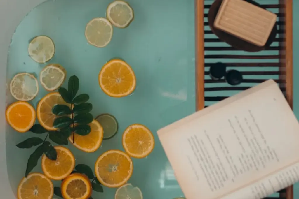 A calming bath scene with slices of lemon and orange floating in turquoise water, a wooden bath tray holding a bar of soap, and an open book for relaxation.