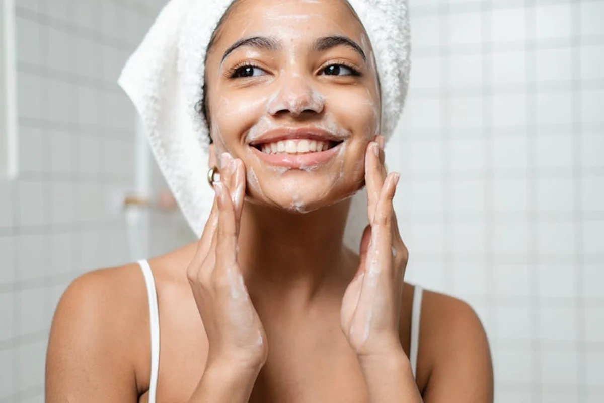 Smiling woman with a towel wrapped around her head, applying foamy face wash in a bathroom.
