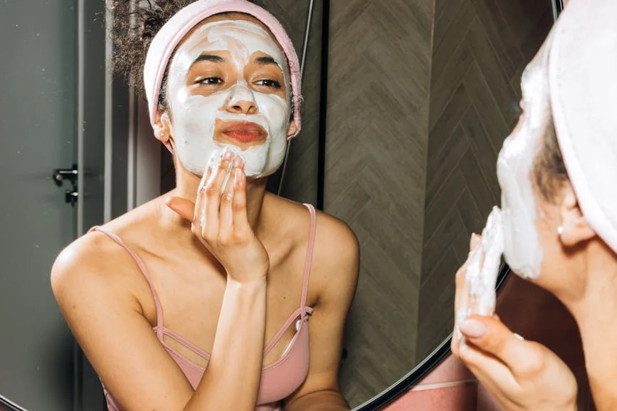Smiling woman with a towel wrapped around her head, applying foamy face wash in a bathroom.